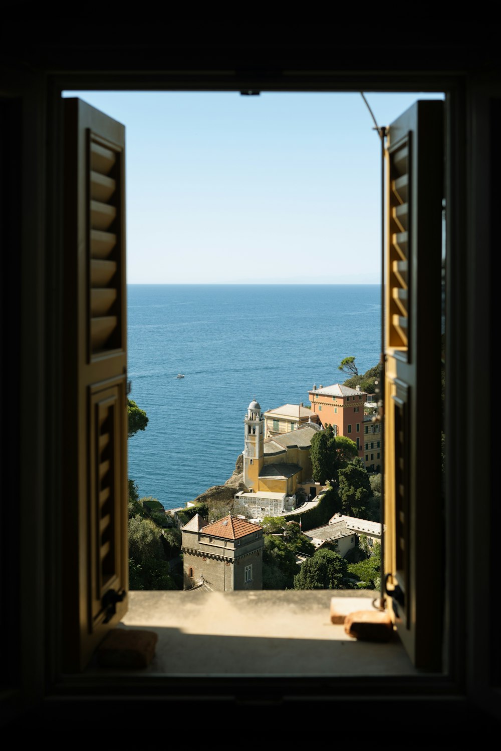 Geöffnetes Fenster mit Holzläden und Blick auf Küstenort und Wasser